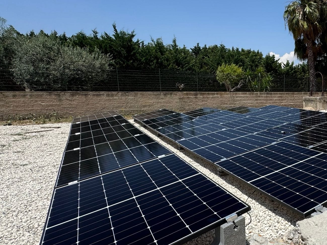 File di pannelli fotovoltaici montati a terra sulla ghiaia, dietro un muro di pietra e una siepe